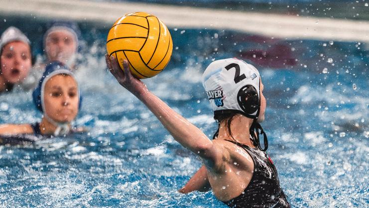 Wasserballspielerin wirft einen gelben Ball im Wasser, während andere Spielerinnen im Hintergrund schwimmen.