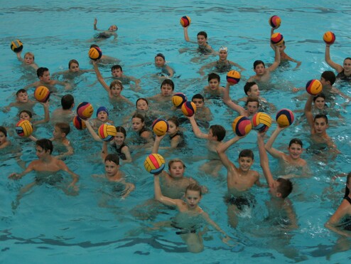 Gruppe von Kindern im Wasser, die Wasserball in den Händen halten und zur Kamera lächeln. Helles, blaues Wasser.
