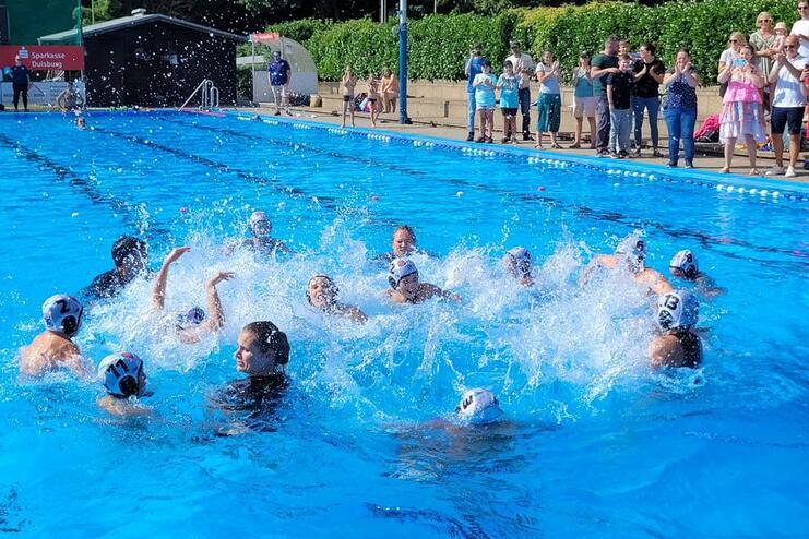 Wasserspiele von mehreren Spielern in einem Schwimmbecken, umgeben von zuschauenden Personen am Beckenrand.