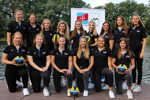 Gruppenfoto eines weiblichen Wasserballteams in schwarzen Trikots mit Wasserb&auml;llen, vor einem See und einem Banner.
