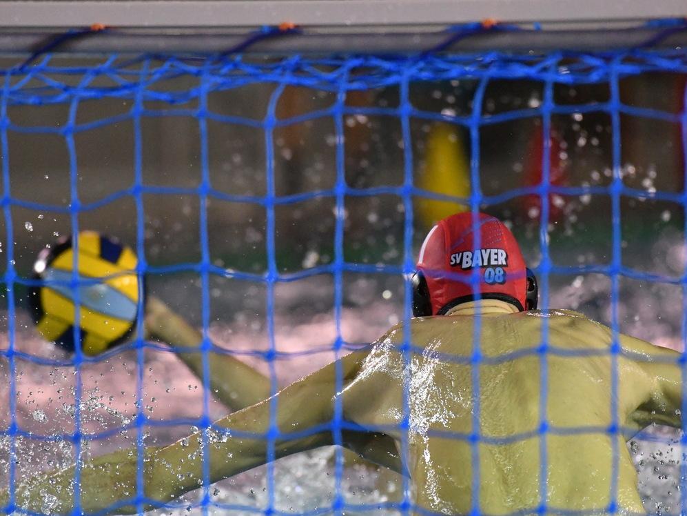 Wasserballspieler in roter Kappe zielt auf das Tor, während Wasser spritzt und der Ball sichtbar ist.