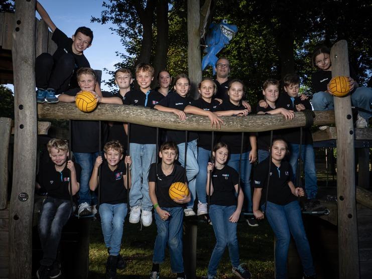 Gruppe von 15 Kindern in schwarzen T-Shirts und einem Erwachsenen auf einem Spielplatz mit Bällen.