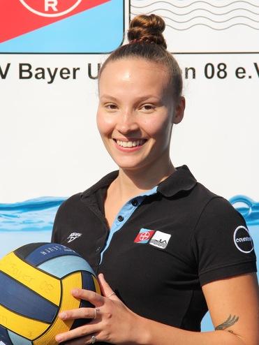 Lächelnde Frau hält einen bunten Wasserball, trägt ein schwarzes Polo, vor einem Hintergrund mit Wasser- und Vereinslogo.