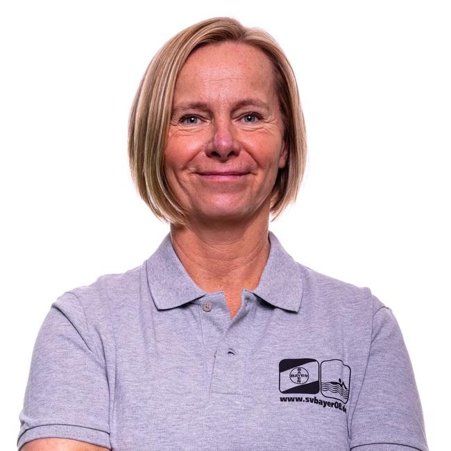 Frau mit kurzem, blondem Haar in grauem Poloshirt, die mit verschränkten Armen leicht lächelt, vor weißem Hintergrund.