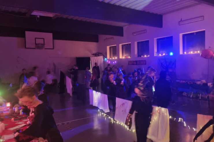 Gemütliche Halloweenfeier in einer Sporthalle mit dekorierten Tischen und kostümierten Gästen in einer festlichen Atmosphäre.