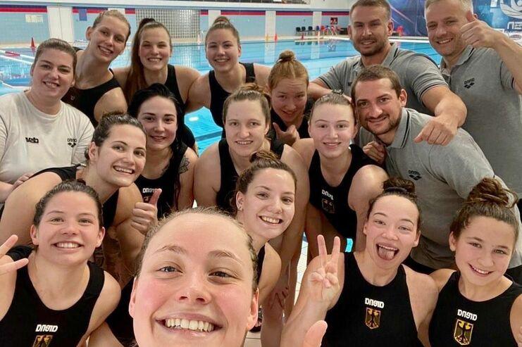 Gruppe von Wasserspringerinnen und Trainern lächelt in einer Sporthalle mit Schwimmbecken im Hintergrund.