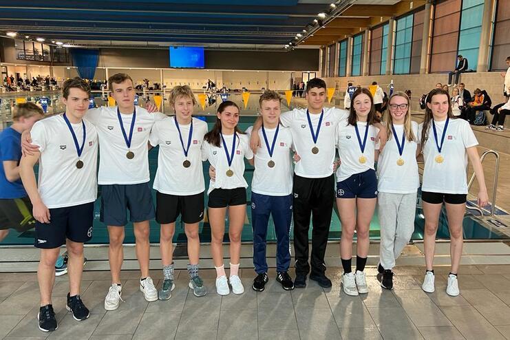 Gruppe von acht Schwimmern in einheitlichen T-Shirts mit Medaillen steht am Beckenrand in einem Schwimmbad.