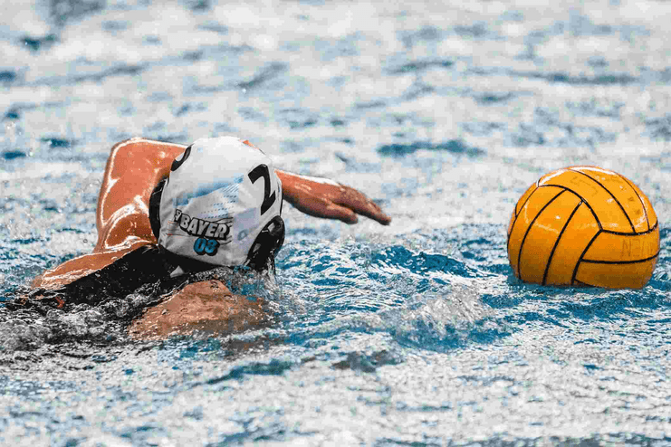 Wasserballspielerin schwimmt mit gelbem Ball im Wasser, Blick nach vorne, weiße Badehaube mit „BAYER 03“.