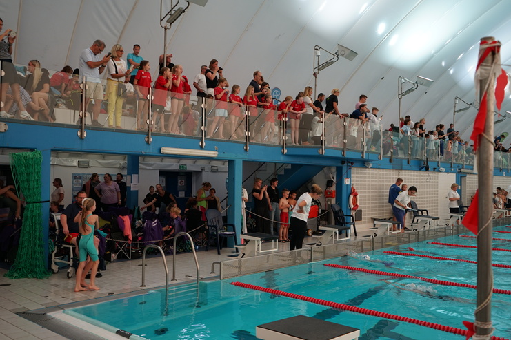 Zuschauer und Schwimmer in einem beheizten Schwimmbad, mit Tribüne und verschiedenen Gruppen am Beckenrand.