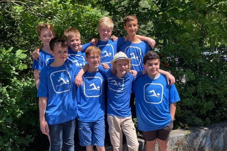 Gruppe von acht Jungen in blauen T-Shirts mit Logo, stehen lächelnd vor einer grünen Alternative mit Holzbank.