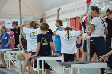 Junge Schwimmer in T-Shirts bereiten sich auf einen Wettbewerb am Schwimmbecken vor, Trainer überwacht die Szene.