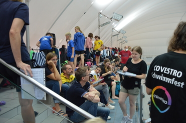 Menschen sitzen und stehen an einer Treppe in einer Schwimmhalle, mit Ausrüstungen und Handzetteln um sie herum.