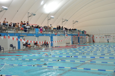 Schwimmbecken mit mehreren Bahnen, Zuschauern auf Balustrade und Schwimmern im Wasser während eines Wettkampfs.