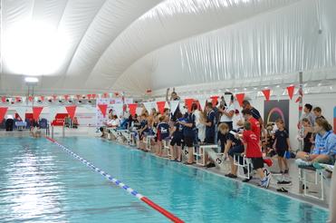 Kinder und Erwachsene stehen am Beckenrand eines Pools, während ein Wettkampf mit rot-weißen Fahnen stattfindet.