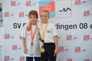 Zwei Jungen in weißen T-Shirts mit Medaillen um den Hals stehen vor einem Banner des SV Bayer.