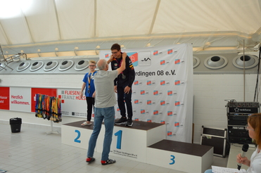 Zwei Schwimmer stehen auf dem Siegertreppchen bei einer Preisverleihung, während ein Dritter eine Medaille erhält.
