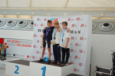 Drei Kinder mit Medaillen stehen auf dem Siegerpodest vor einer Bannerwand bei einer Sportveranstaltung.