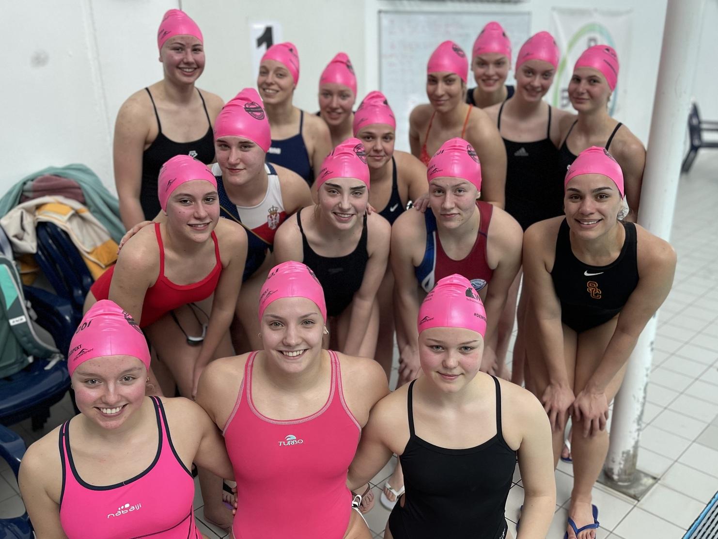 Gruppe von 16 Schwimmerinnen in pinken Badekappen, posierend am Beckenrand in einem Schwimmhallenraum.
