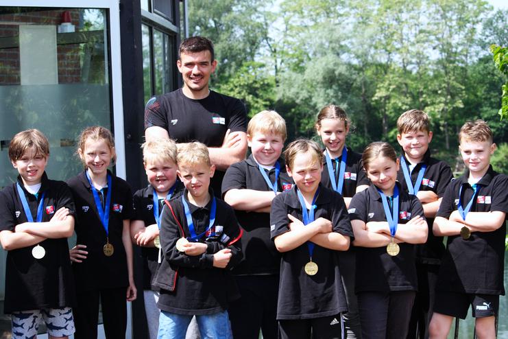Gruppe von Kindern mit Medaillen, stehend vor einem Geb&auml;ude, l&auml;chelnd und in schwarzen T-Shirts.