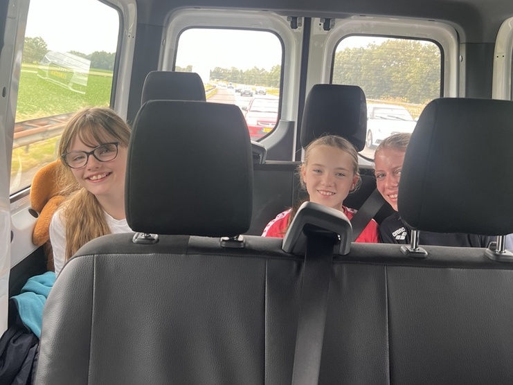 Drei lächelnde Kinder sitzen im Rücksitz eines Fahrzeugs, umgeben von Autositzen und einem Fensterblick auf die Landschaft.