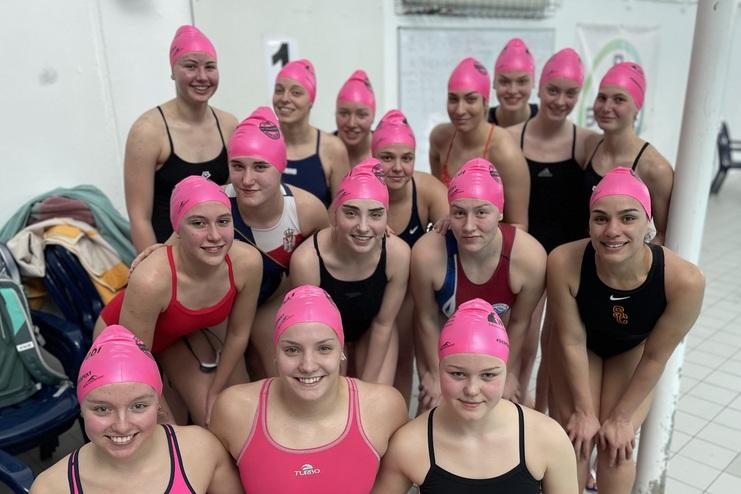 Gruppe von 16 Schwimmerinnen in pinken Badekappen und Badeanzügen, posierend am Beckenrand in einem Schwimmbad.