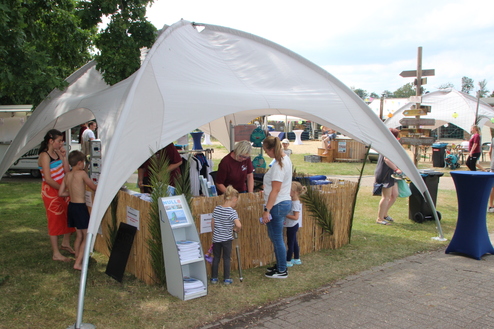 Wettbewerb mit Zelt, Leuten und Ständen auf einer Wiese, umgeben von grünen Bäumen und einem entspannten Veranstaltungsambiente.