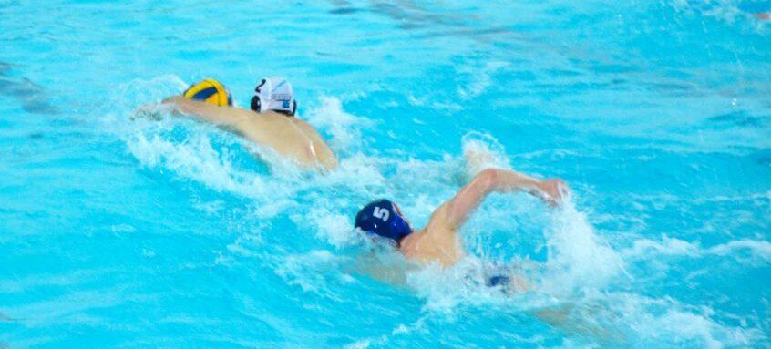 Zwei Wasserballspieler schwimmen in einem blauen Schwimmbecken, einer mit gelbem Ball, der andere mit blauer Kappe.