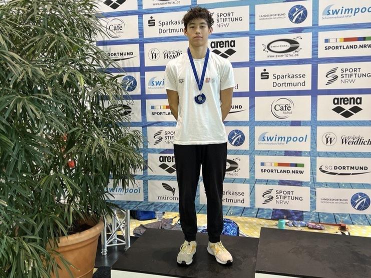 Männlicher Schwimmer mit Medaille steht auf dem zweiten Platz einer Siegerehrung vor Werbebannern und einer Pflanze.