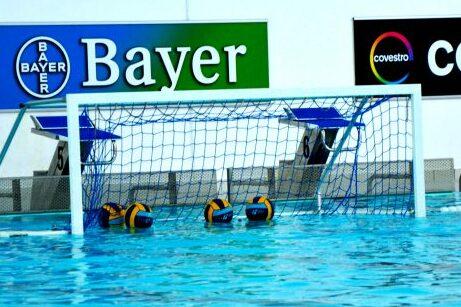 Waterpolo-Tor mit Bällen im Wasser, umgeben von blauer Wasseroberfläche und Sponsorenlogos im Hintergrund.