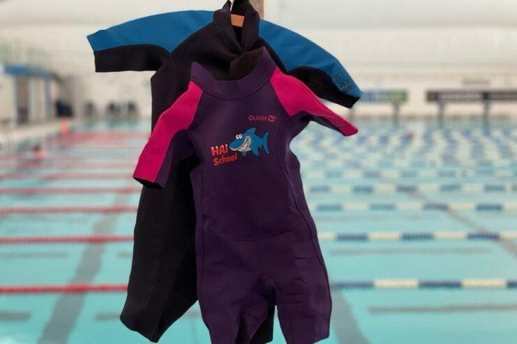 Drei Schwimmanzüge hängen an einem Ständer vor einem Schwimmbecken mit blauen Bahnen.