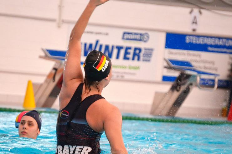 Wasserballspielerin hebt die Hand in einem Schwimmbecken; eine weitere Spielerin schaut im Hintergrund.