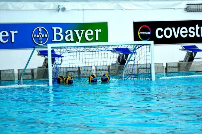 Waterpolo-Tor mit Bällen im Wasser, umgeben von blauer Wasseroberfläche und Sponsorenlogos im Hintergrund.