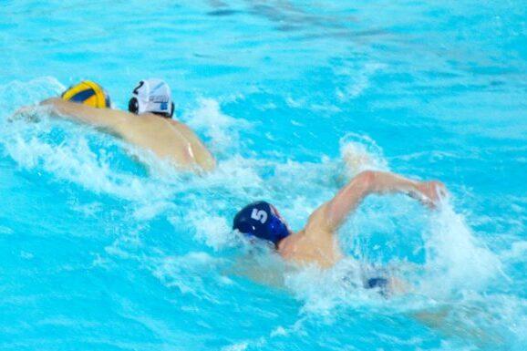 Zwei Wasserballspieler schwimmen in einem blauen Schwimmbecken, einer mit gelbem Ball, der andere mit blauer Kappe.