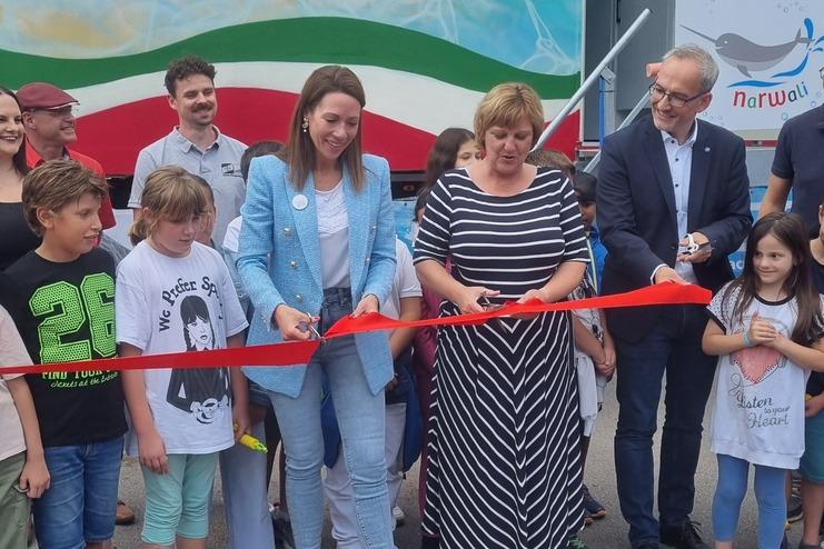 Eröffnungsevent mit mehreren Personen, die ein rotes Band schneiden, umgeben von Kindern und einem bunten Hintergrund.