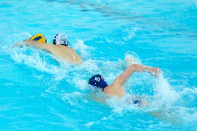 Zwei Wasserballspieler schwimmen in einem blauen Schwimmbecken, einer mit gelbem Ball, der andere mit blauer Kappe.