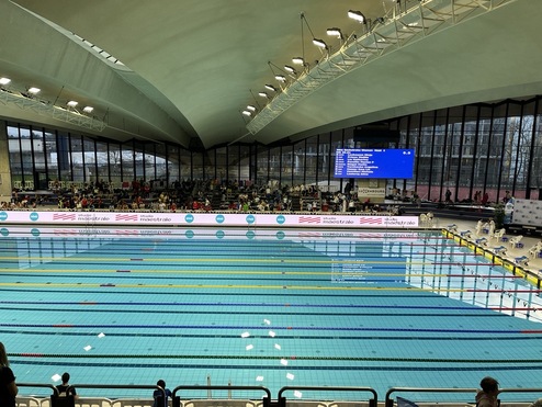 Schwimmbecken in einer modernen Halle, gefüllt mit klarem Wasser und bunten Bahnmarkierungen, Zuschauer sitzen im Hintergrund.