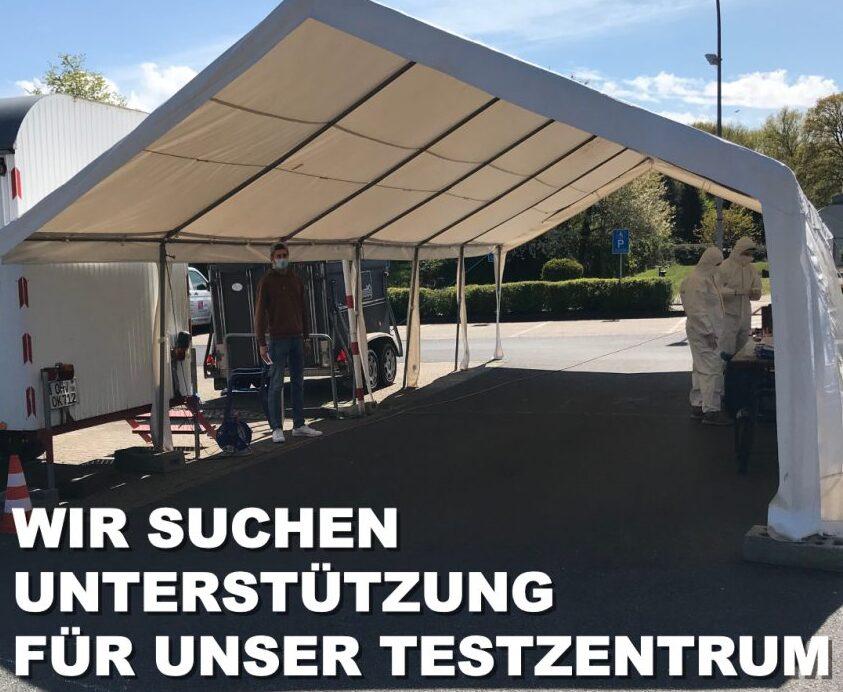 Zelt mit Personen in Schutzanzügen, ein Mann steht daneben; Beschilderung zur Unterstützung für ein Testzentrum.