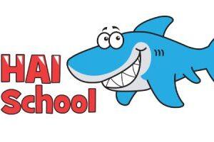 Grafik eines l&auml;chelnden blauen Haifischs neben dem Schriftzug &bdquo;HAI School&ldquo; in roter Schrift.