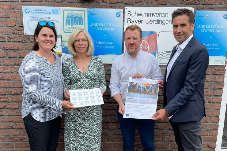 Vier Personen stehen vor einer Backsteinwand, halten Unterlagen und ein Plakat des Schwimmvereins Bayer Uerdingen.
