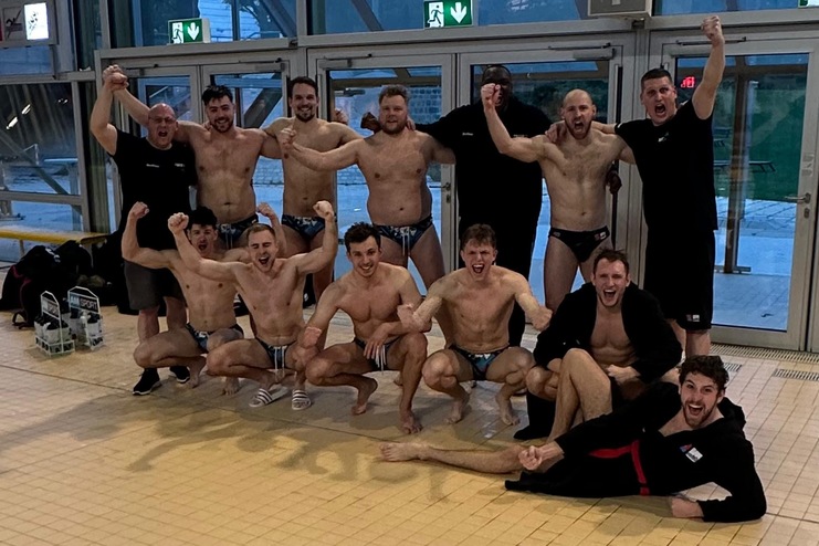 Gruppe von Männern in Badebekleidung posiert fröhlich in einem Schwimmbad mit großen Fenstern.
