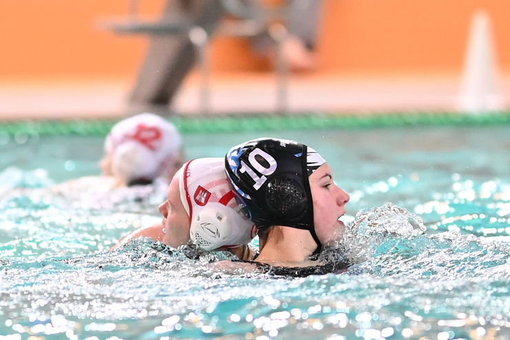 Zwei Wasserballspielerinnen in einem Wettkampf schwimmen gegeneinander, mit spritzendem Wasser im Hintergrund.