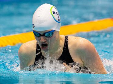Sportlerin im schwarzen Badeanzug schwimmt im Wettkampfmodus, Spritzer umgeben sie im Wasser, mit einer Badekappe und Brille.