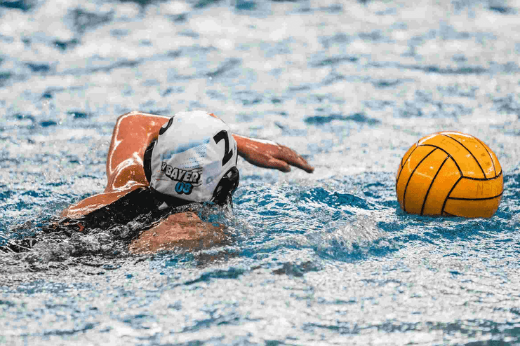 Wasserballspielerin schwimmt mit gelbem Ball im Wasser, Blick nach vorne, weiße Badehaube mit „BAYER 03“.