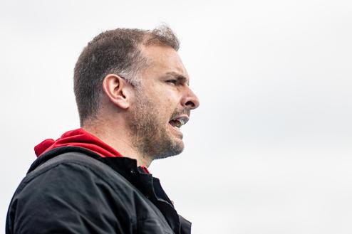 Mann spricht mit leidenschaftlichem Ausdruck, im Profil vor einem grauen Himmel. Er trägt eine schwarze Jacke mit rotem Hoodie.