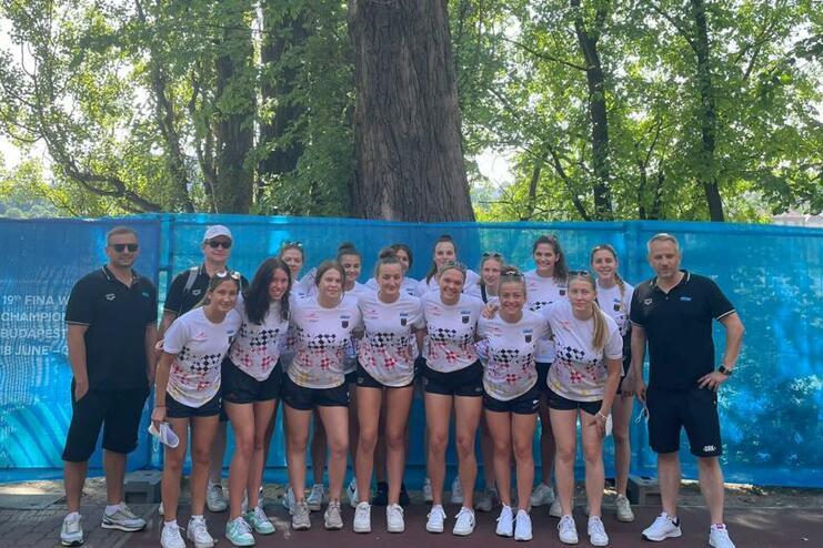 Gruppe von zwölf Frauen in Sportbekleidung, Posieren vor einem blauen Banner und großen Bäumen im Hintergrund.