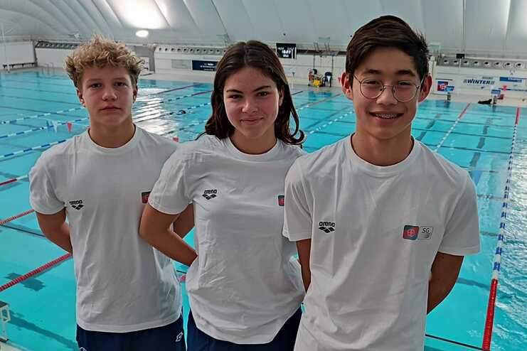Drei Jugendliche in weißen T-Shirts stehen vor einem Schwimmbecken mit blauer Wasseroberfläche und roten Fähnchen.