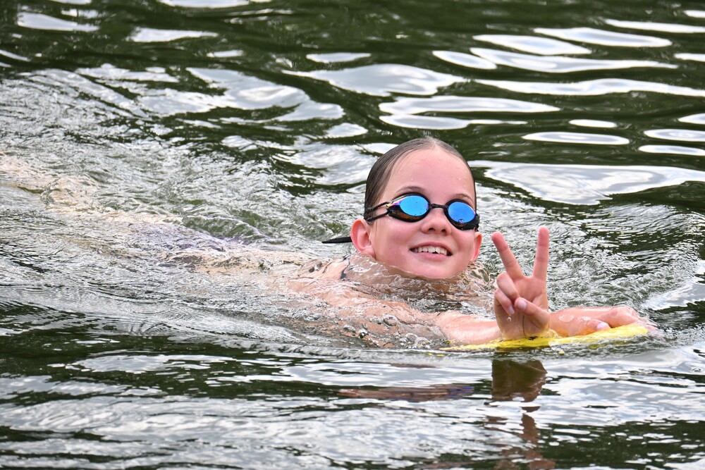Ein Mädchen mit Schwimmbrille schwimmt lächelnd im Wasser und zeigt das Friedenszeichen mit einer Hand.