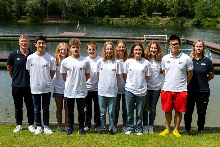 Gruppenfoto von sechs Jugendlichen und drei Trainern am Ufer eines Gewässers, umgeben von Bäumen und Bootsstegen.