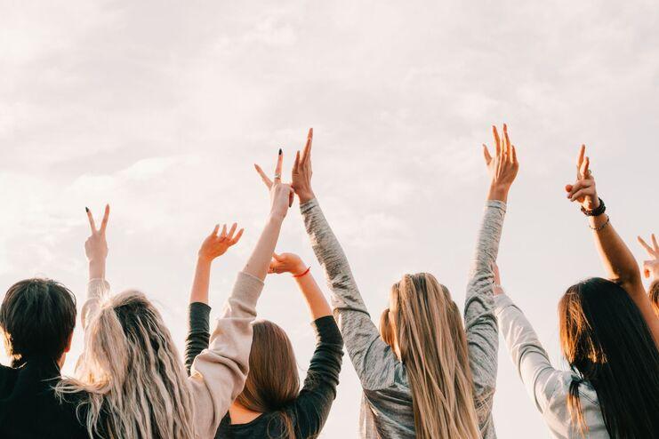 H&auml;nde von mehreren Personen heben sich zum Himmel, w&auml;hrend sie eine friedliche Geste machen, vor einem hellen Wolkenhimmel.