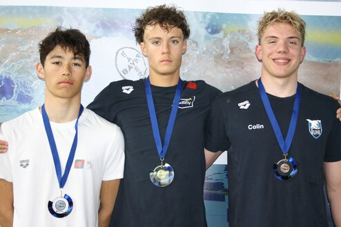 Drei Medaillengewinner stehen vor einem Hintergrund mit Wasser, jeder trägt eine Medaille und blickt in die Kamera.